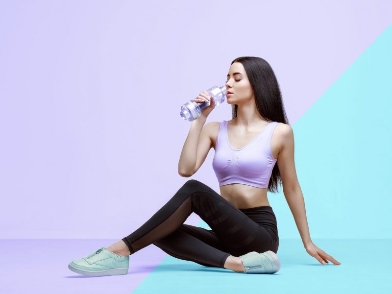Amazing woman in trendy sportswear drinking water from bottle to stay hydrated after workout. Beautiful slim brunette young girl in fashion leggings and purple top. Fitness, healthy lifestyle, sport.